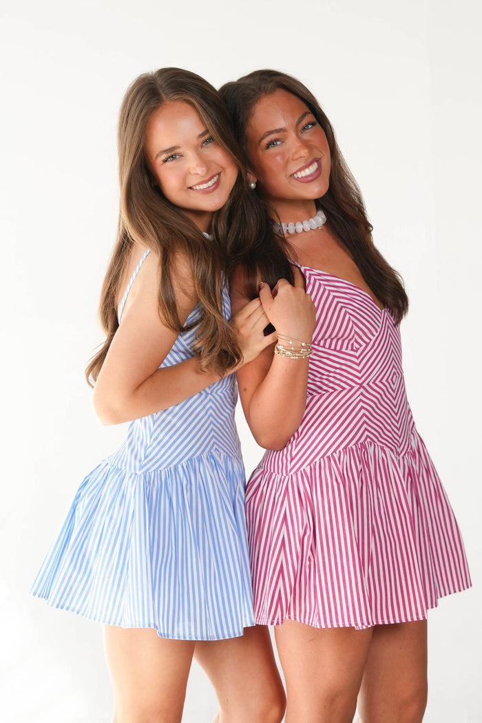 Two young women posing together against a white studio background, each wearing a short pleated dress with vertical stripes. The woman on the left wears a light blue and white striped dress, and the woman on the right wears a pink and white striped dress.