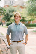 A smiling young man standing outdoors on a campus walkway, wearing a light blue polo shirt with a small embroidered logo on the left chest, khaki shorts, and a brown belt.