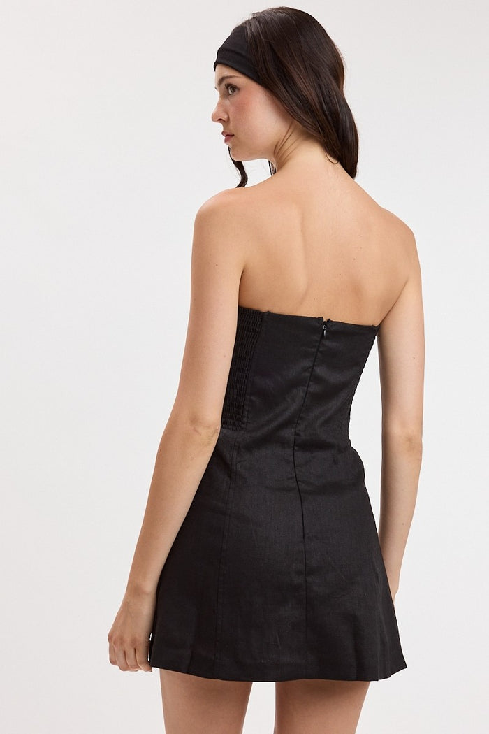 A woman shown from the back wearing a strapless black mini dress with a fitted bodice and flared skirt, photographed against a plain light gray studio background.