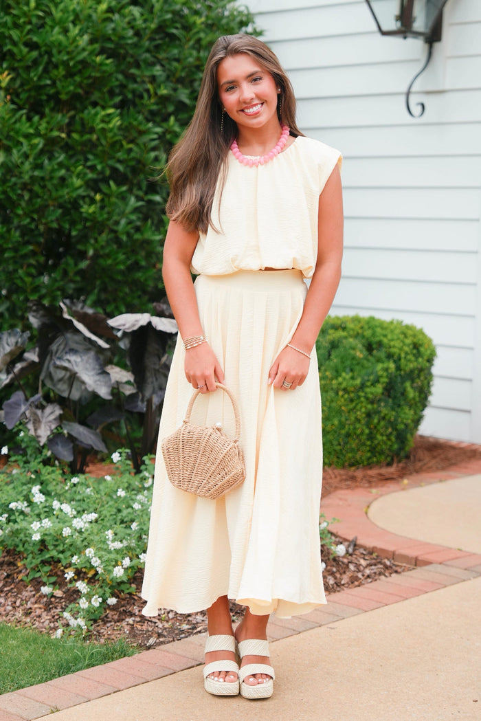Woman in a light yellow dress holding a straw bag outdoors.
