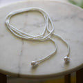 A delicate silver beaded necklace with two large teardrop-shaped silver beads at the ends, displayed on a light marble surface.