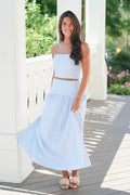 Woman wearing a light blue dress standing on a wooden deck with greenery in the background