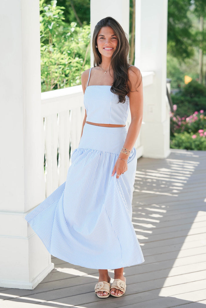 Woman wearing a light blue dress standing on a wooden deck with greenery in the background