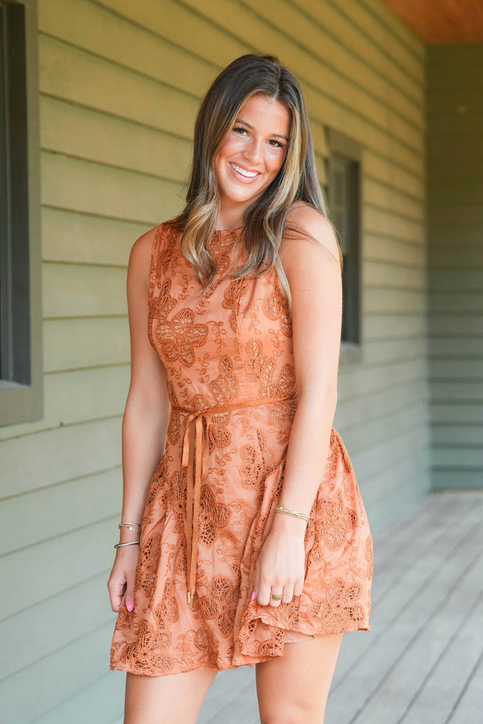 A woman wearing a sleeveless, rust-orange lace dress with a tie waist, standing on a porch with light green house siding in the background.