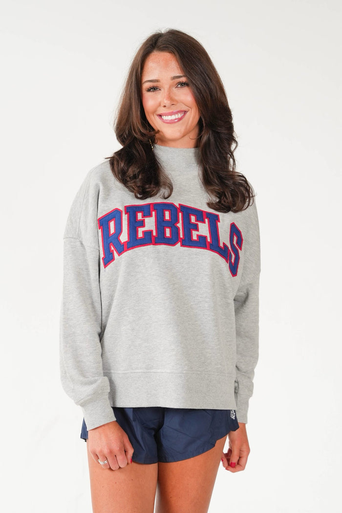 A woman wearing a light gray crewneck sweatshirt with 'REBELS' printed across the chest in red and blue varsity lettering, paired with navy blue shorts, against a plain white studio background.