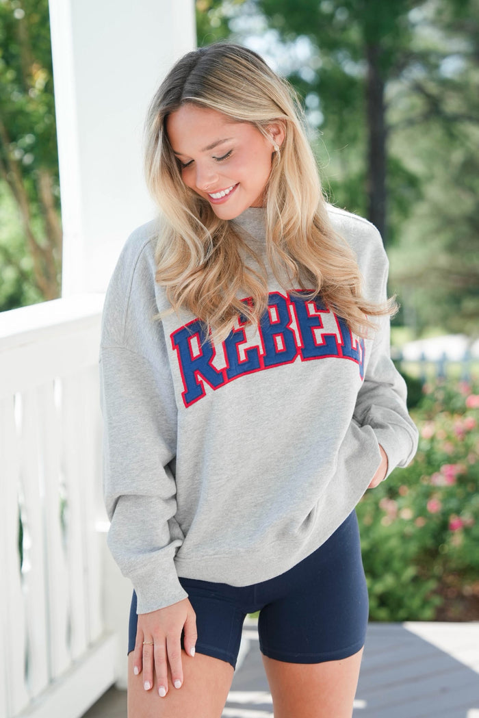 Woman wearing a gray sweatshirt with 'REBEL' printed on it, standing outdoors.