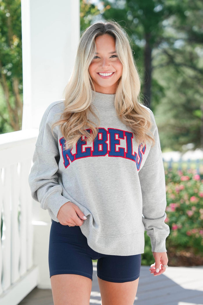 Woman wearing a gray sweatshirt with 'REBEL' printed on it, standing outdoors.