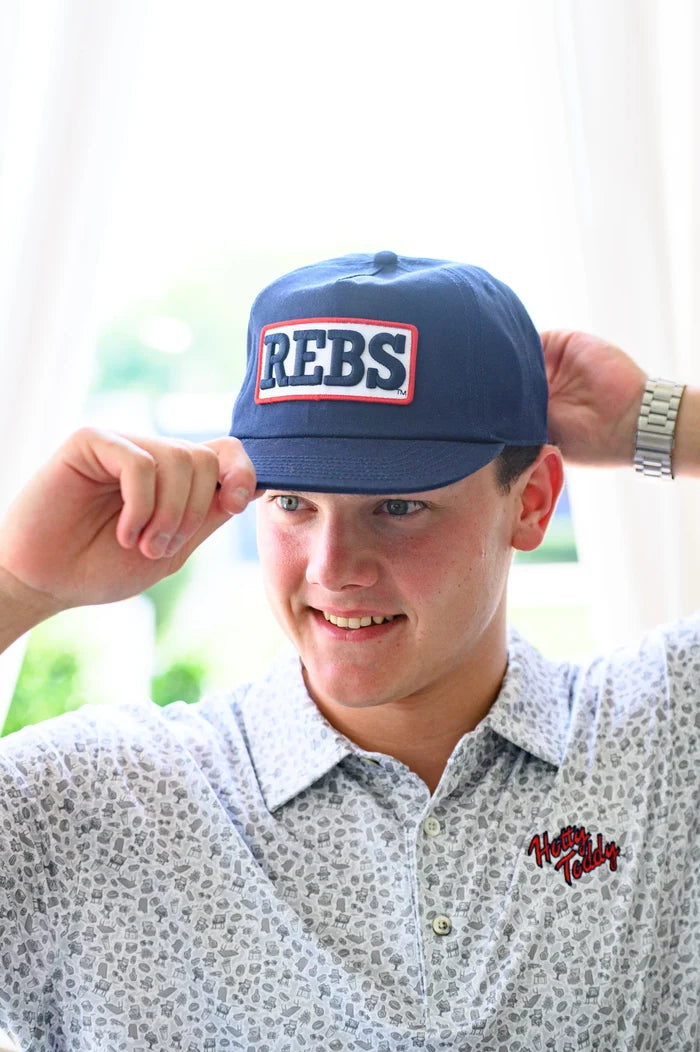 A smiling young man wearing a navy blue baseball cap with 'REBS' embroidered in white and red, paired with a light patterned button-down shirt.