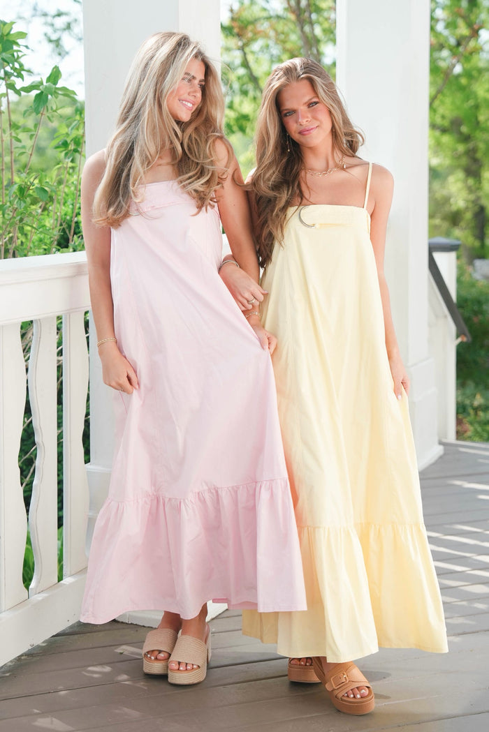 Two women wearing light pink and yellow dresses standing on a wooden deck with greenery in the background.