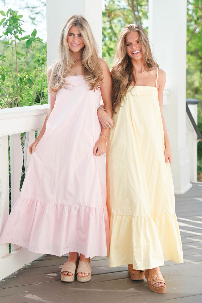 Two women wearing long dresses standing on a wooden deck with greenery in the background.
