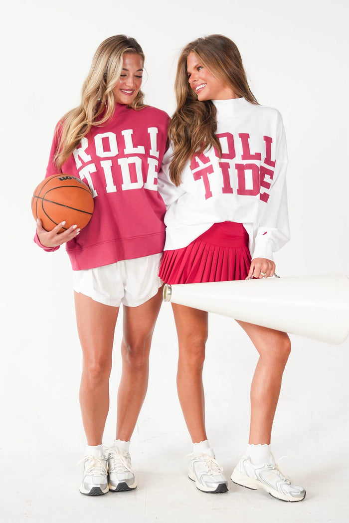 Two women modeling matching 'ROLL TIDE' sweatshirts in a studio setting. The woman on the left wears a dark pink sweatshirt and holds an orange basketball, while the woman on the right wears a white sweatshirt showing the back with the same red text and a red pleated skirt.