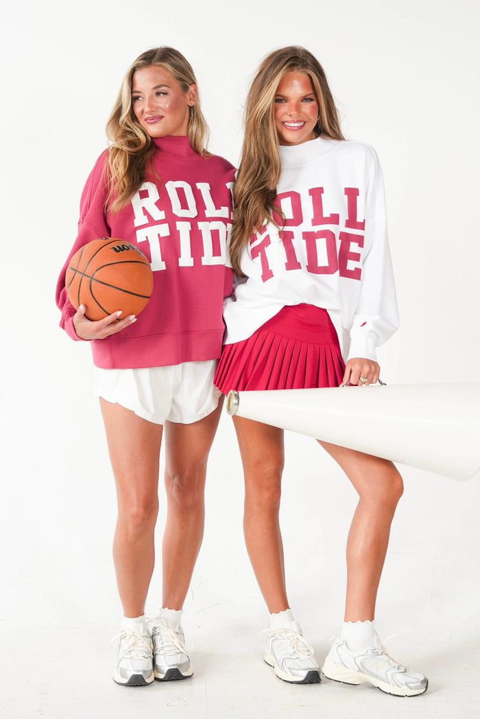 Two women modeling matching athletic sweatshirts with 'ROLL TIDE' printed in red. The woman on the left wears a magenta sweatshirt and holds a basketball, while the woman on the right wears a white sweatshirt and a red pleated skirt.