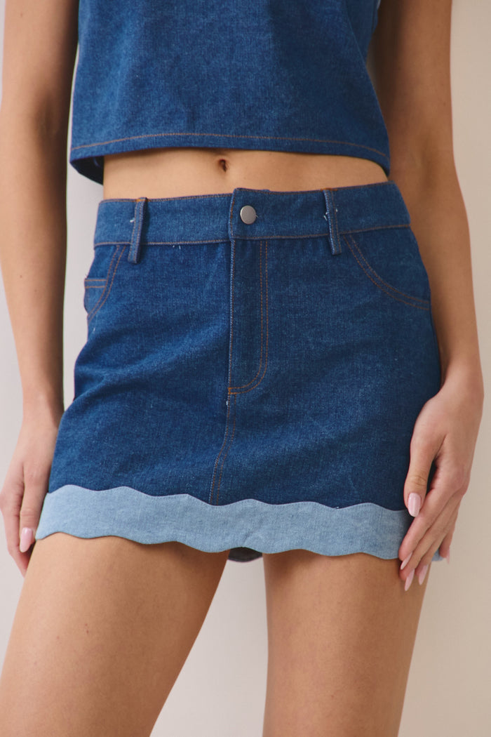 Model posing in a dark blue denim skirt with a light blue scalloped hem in front of a beige wall.