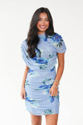 A woman modeling a light blue ruched mini dress with a blue and green floral print, set against a plain white studio background.
