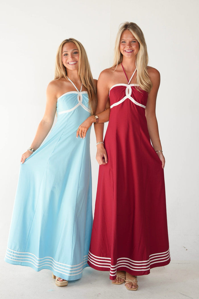 Two women modeling halter maxi dresses side by side against a plain white studio background. The woman on the left wears a light blue dress with white trim, and the woman on the right wears a dark red dress with white trim.