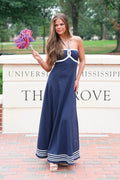 A woman stands outdoors in front of a University of Mississippi sign, wearing a navy blue strapless maxi dress with white trim and striped detailing at the hem. She is holding a colorful pom-pom.