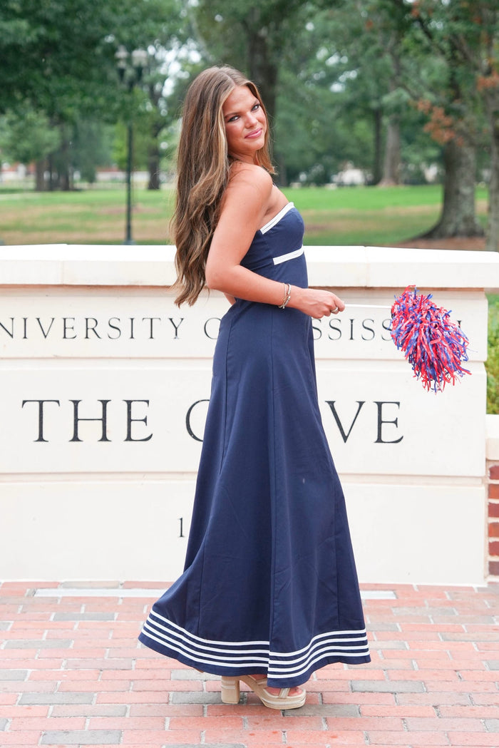 A woman modeling a navy blue strapless maxi dress with white stripe accents, standing outdoors in front of a white brick sign that reads 'UNIVERSITY OF GEORGIA THE COLLEGE'. She is holding a red and blue pom-pom.