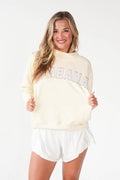 A woman modeling a cream-colored Alabama sweatshirt with 'ALABAMA' printed across the chest in gray and white, paired with white shorts, against a plain white background.