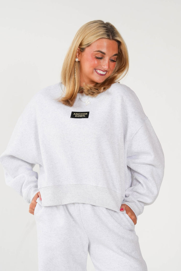 A woman wearing a light gray heather sweatshirt and matching sweatpants, featuring a small black rectangular chest patch with white text that reads 'PRAIRIE DOGS'.