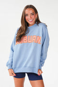 A woman wearing a light blue crewneck sweatshirt with 'AUBURN' in orange and white gingham lettering across the chest, paired with navy blue bike shorts, against a plain white studio background.