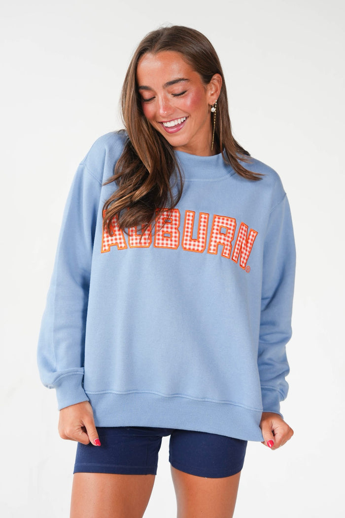 A woman wearing a light blue crewneck sweatshirt with 'AUBURN' printed across the chest in red and white gingham letters, paired with navy blue bike shorts, against a plain white background.