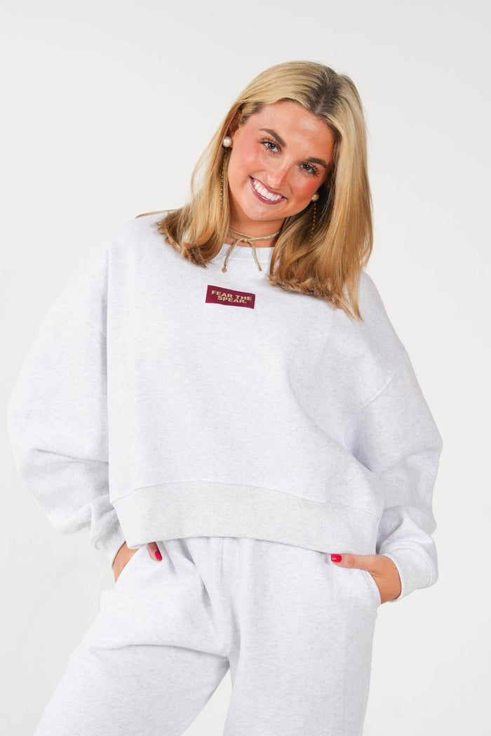 A woman wearing a light gray heather sweatshirt and matching sweatpants, featuring a small red rectangular logo patch on the chest that reads 'Supreme' in white text, set against a plain light gray background.
