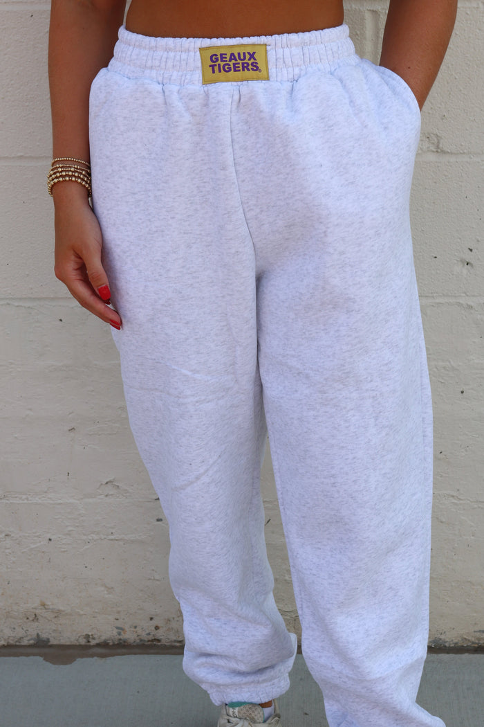 A person wearing light heather gray sweatpants with a gold 'Beaux Tigers' patch on the waistband, standing against a white painted brick wall.