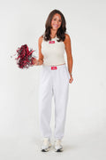 A woman modeling a white sleeveless crop top and matching white sweatpants, holding a red and black pom-pom against a plain white studio background.