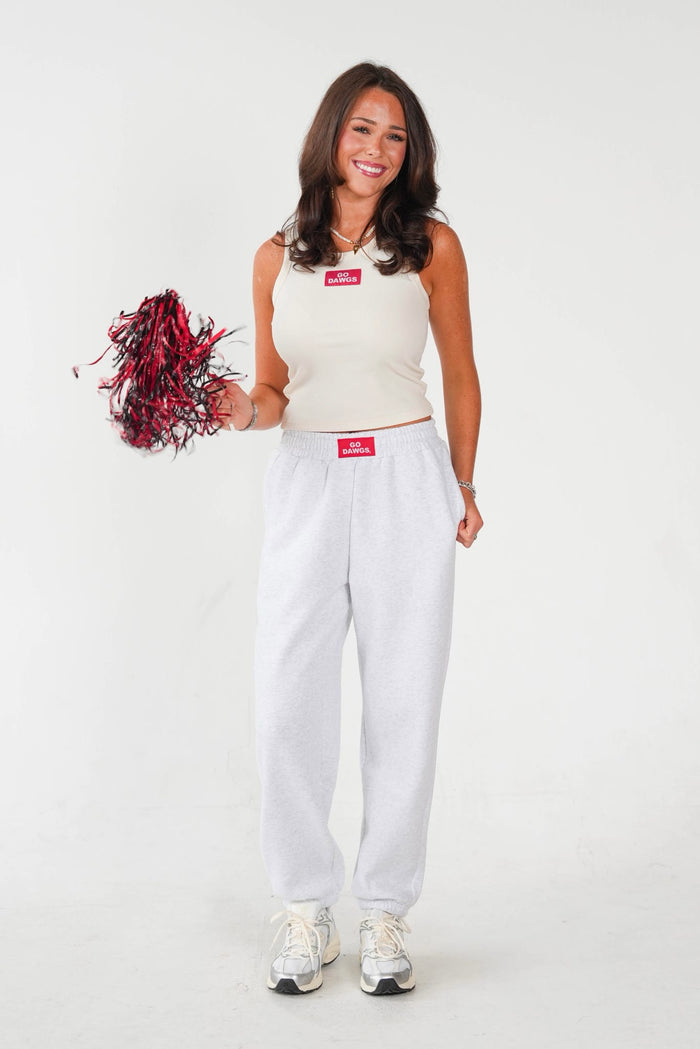 A woman modeling a white sleeveless crop top and matching white sweatpants, holding a red and black pom-pom against a plain white studio background.