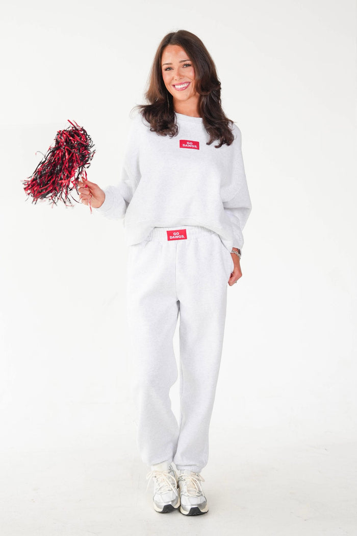 A woman modeling a white sweatshirt and sweatpants set with red Supreme box logos, holding a red and black pom-pom against a plain white background.