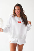 A woman modeling a light heather gray sweatshirt and matching shorts set, featuring orange rectangular logo patches on the chest and shorts.