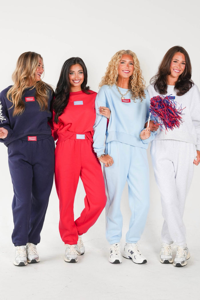 Four women modeling matching tracksuits in navy, red, light blue, and white, each with a small red rectangular patch on the chest, standing against a plain white studio background.
