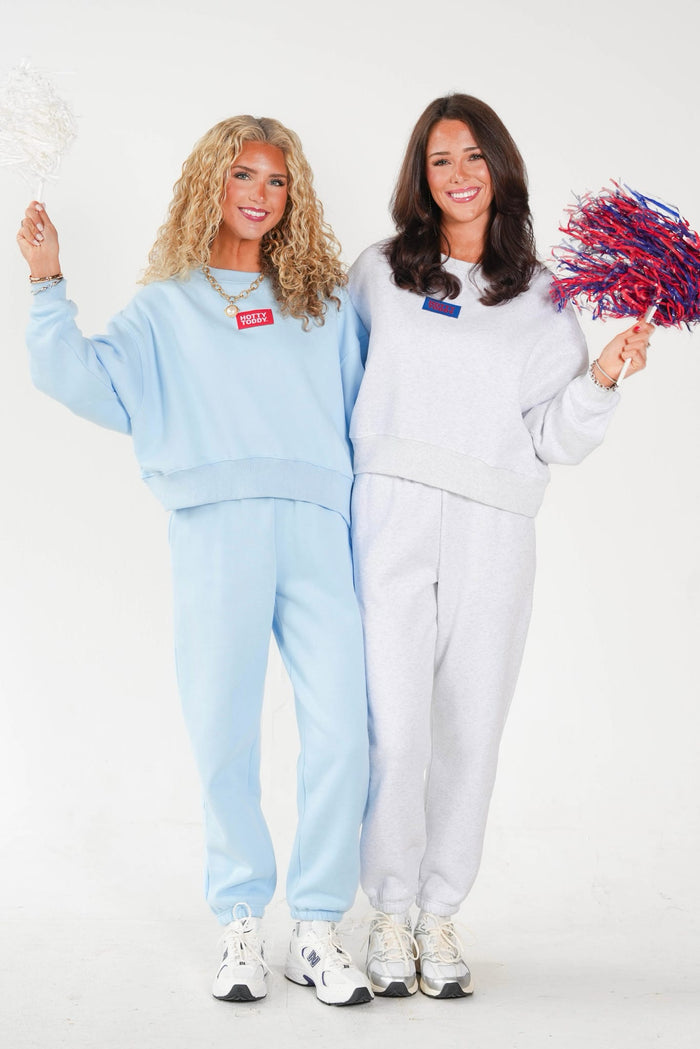 Two women modeling matching sweatshirt and jogger sets, standing against a plain white studio background. The woman on the left wears a light blue set and holds a white pom-pom, while the woman on the right wears a light gray set and holds a red and blue pom-pom.