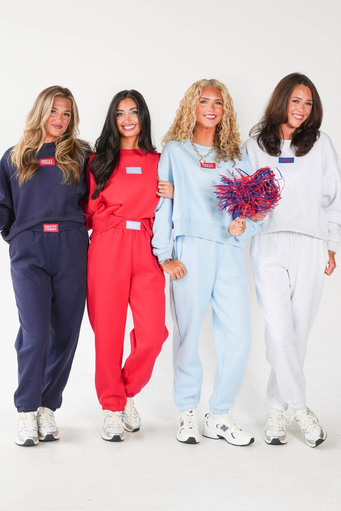 Four women modeling matching sweatshirt and jogger sets in navy, red, light blue, and white, each with a small rectangular chest patch. One woman holds a red and blue pom-pom against a plain white studio background.