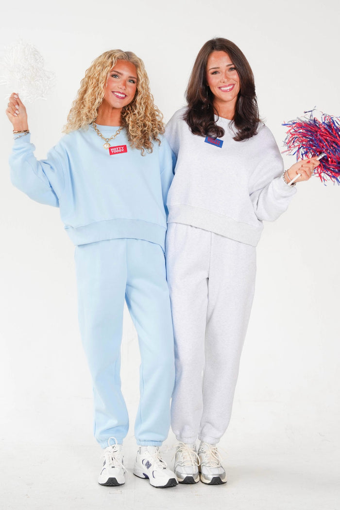 Two women modeling matching sweatshirt and jogger sets, one in light blue and one in light gray, each with a small red rectangular chest patch. They are standing against a plain white studio background, smiling and holding white and red/blue pom-poms.