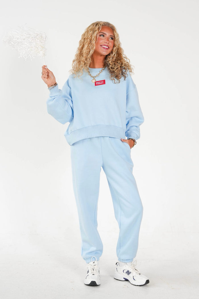 A woman modeling a light blue sweatshirt and matching jogger pants set, featuring a red rectangular chest patch with white text, paired with white sneakers, against a plain white studio background.