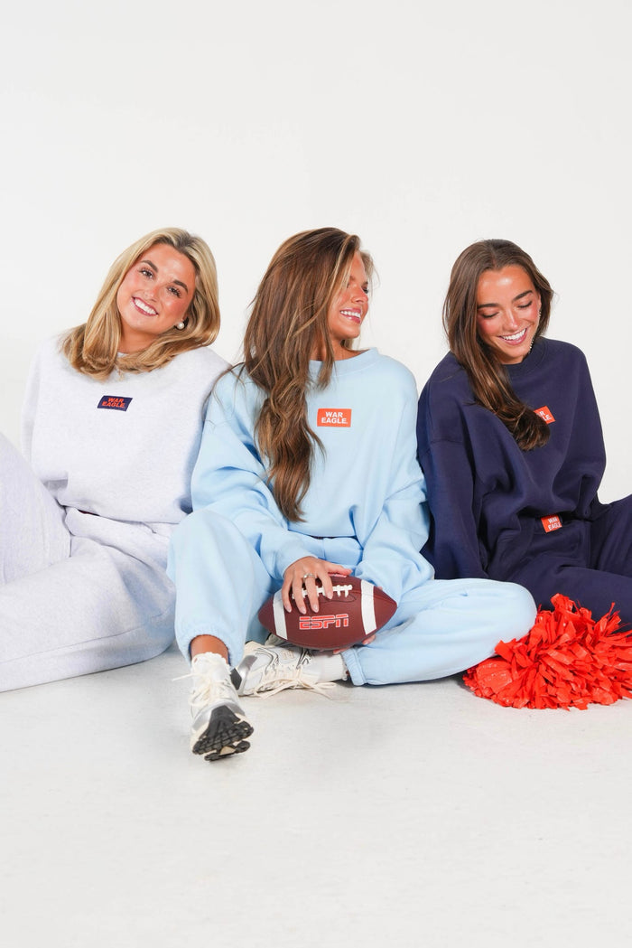 Three women sitting on a white studio floor wearing matching oversized sweatpants and sweatshirts in white, light blue, and navy, each with a small red rectangular chest patch. The center woman holds a brown football, and red pom-poms sit to the right.