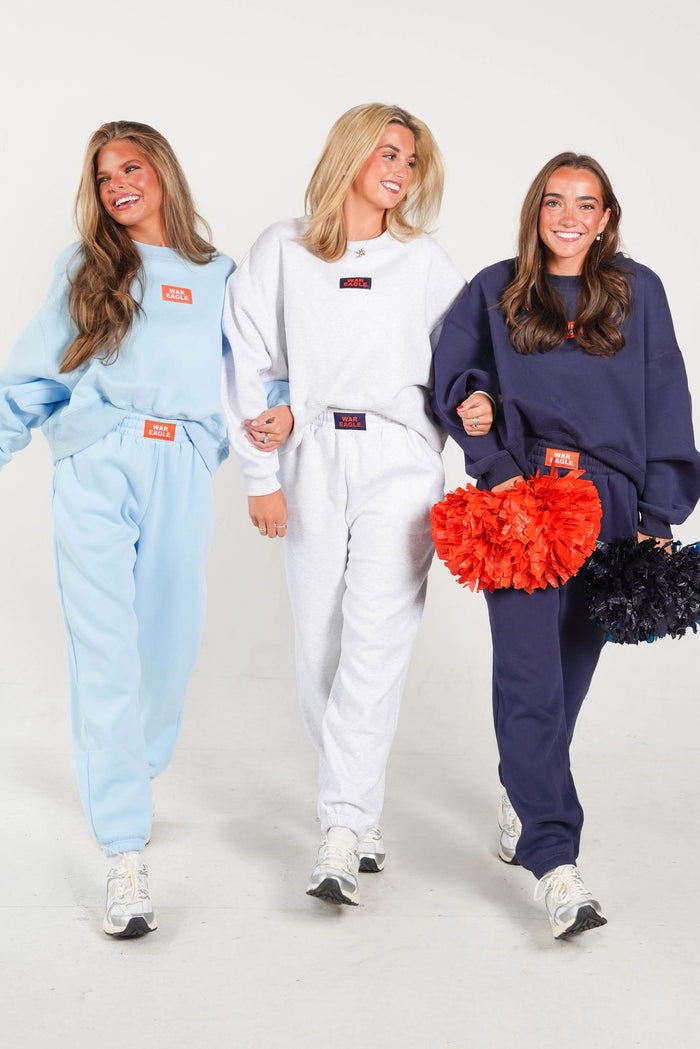 Three women walking side-by-side in matching oversized sweatshirt and jogger tracksuits. The left woman wears a light blue set, the middle woman wears a white set, and the right woman wears a navy blue set.