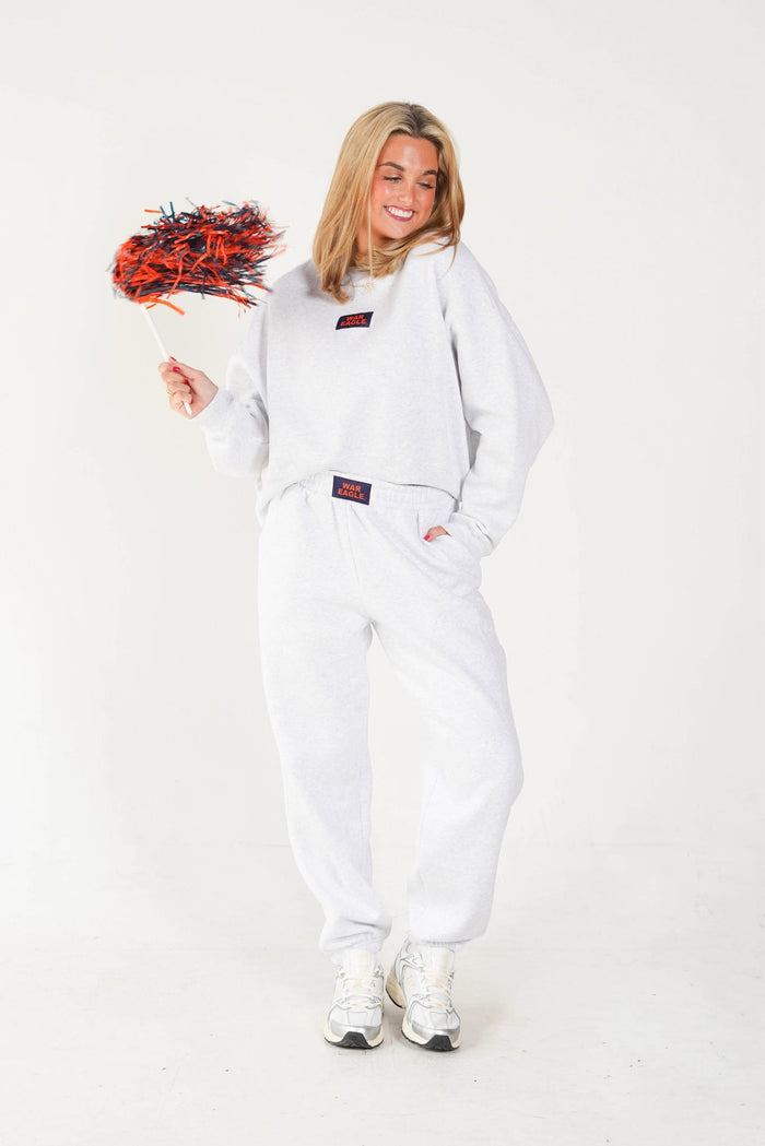 A woman modeling a white sweatshirt and matching sweatpants set, holding a small red pom-pom against a plain white studio background.