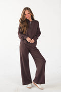 A woman modeling a matching dark brown lounge set consisting of a long-sleeve half-zip pullover and wide-leg pants, paired with white sneakers, against a plain white studio background.