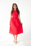 A woman modeling a bright red sleeveless midi dress with a high neckline and gathered waist, paired with white platform sandals and a pearl necklace, against a plain white studio background.