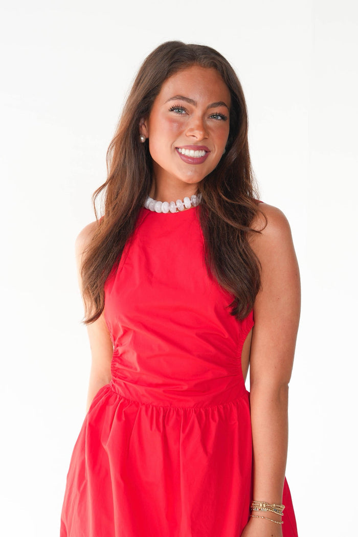 A smiling woman with long brown hair wearing a bright red sleeveless dress and a white pearl necklace, standing against a plain white background.