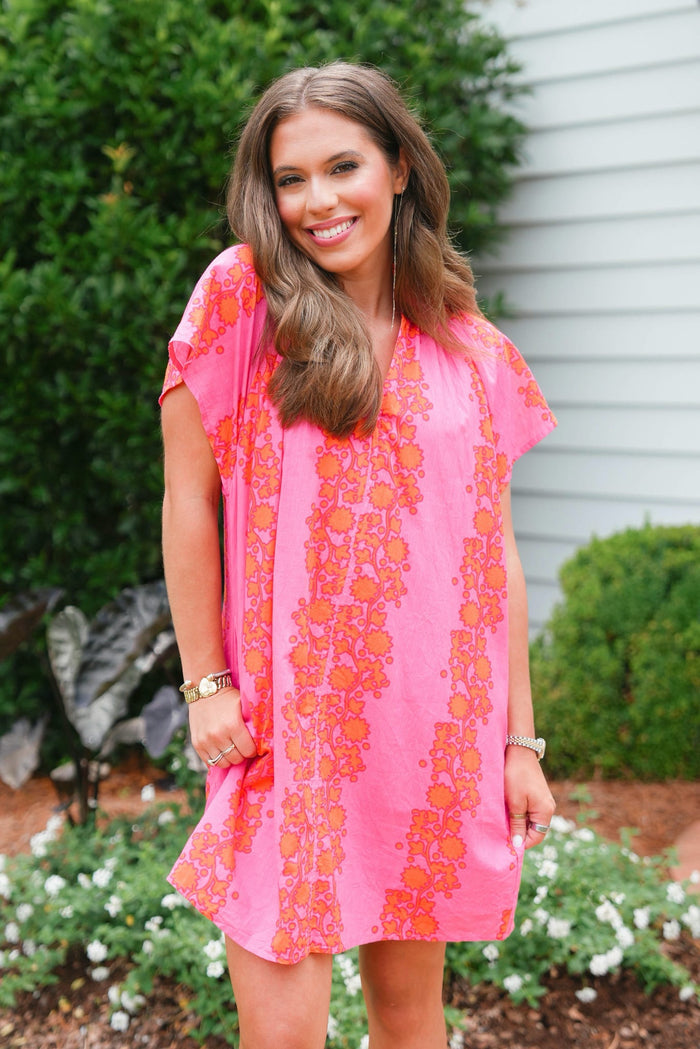 Woman wearing a pink dress with orange floral pattern standing outdoors.
