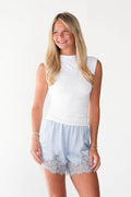 A woman modeling a white sleeveless mock-neck top and light blue shorts with a lace hem, set against a plain white studio background.