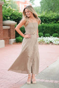 A woman modeling a sleeveless taupe maxi dress with a buttoned bodice and a large round belt buckle, standing outdoors on a brick walkway with green hedges in the background.