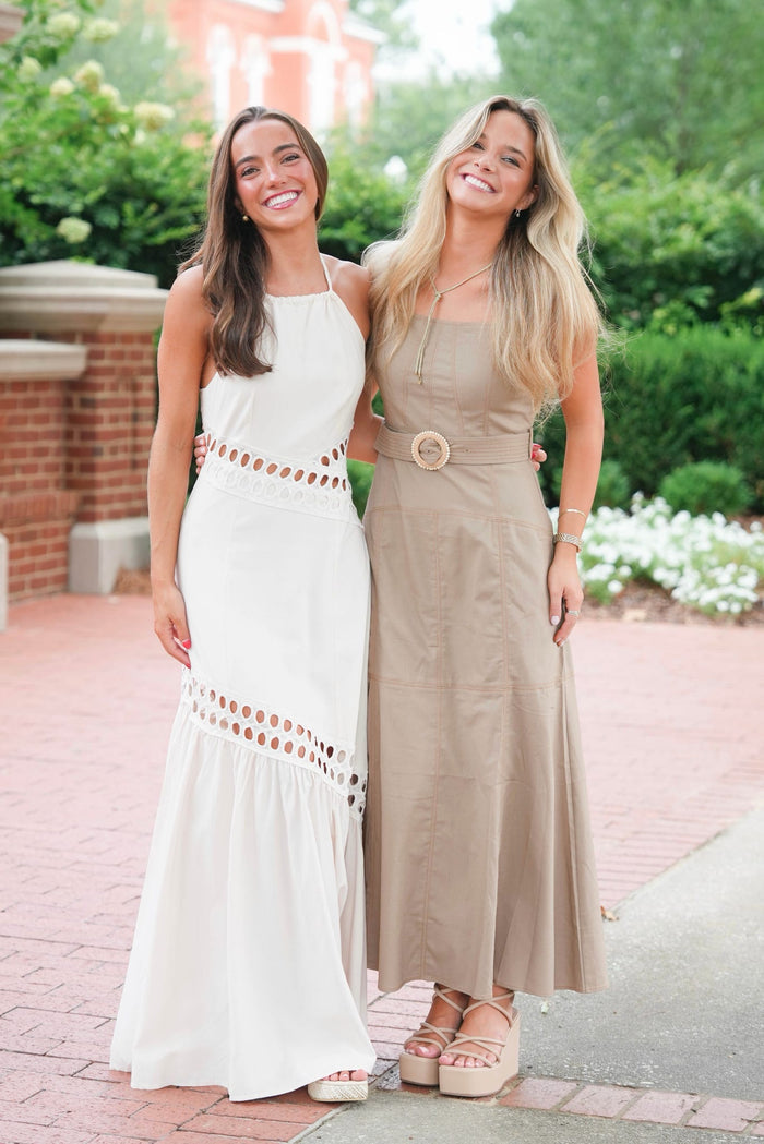 Two women standing outdoors on a brick walkway wearing long sleeveless maxi dresses, one in white with brown cutout details and one in taupe with a circular belt detail.