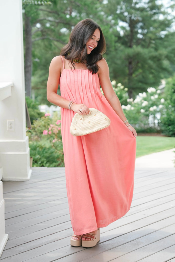 Woman in a coral dress holding a white handbag on a wooden deck with greenery in the background