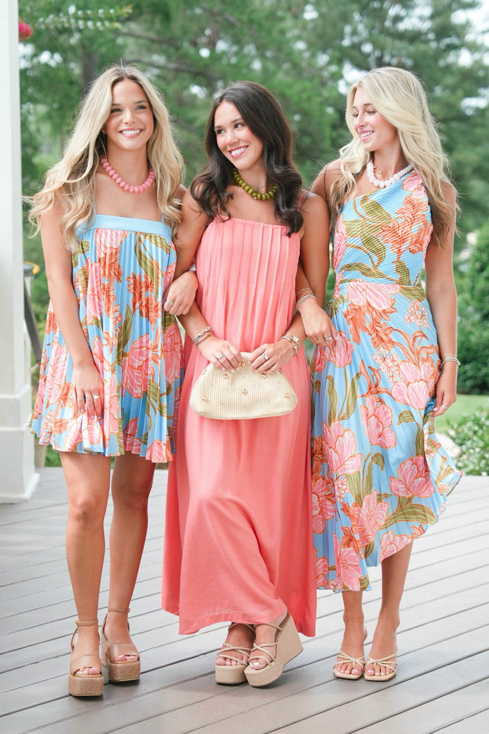 Three women in floral dresses standing on a wooden deck with greenery in the background