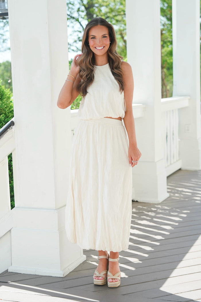 Woman in a white dress standing on a wooden deck with greenery in the background