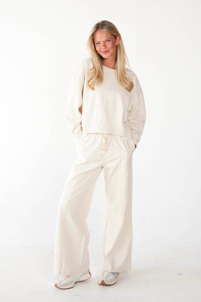 A woman modeling a matching cream-colored lounge set consisting of a long-sleeve sweatshirt and wide-leg drawstring pants, paired with white sneakers, against a plain white studio background.